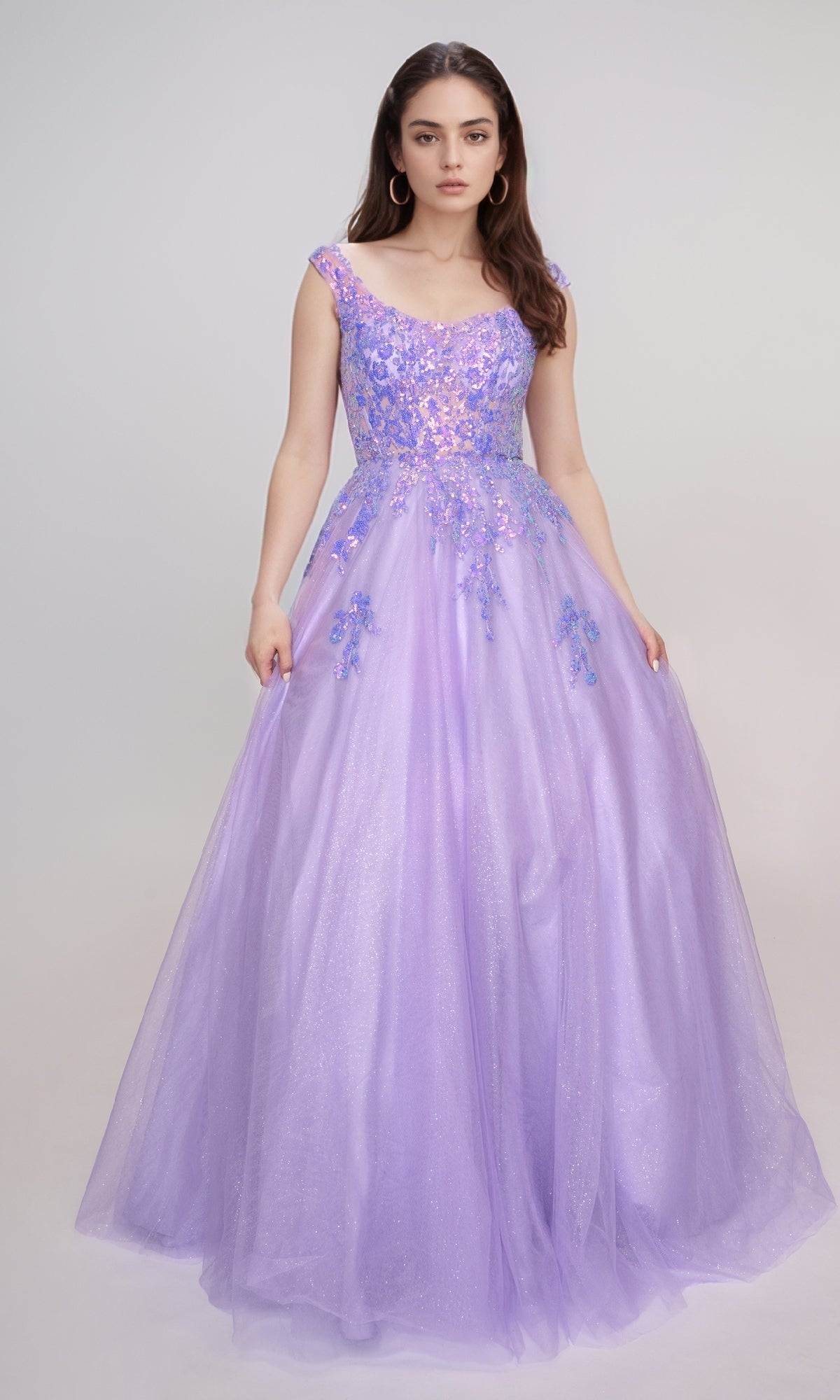 A woman stands against a plain background in a lavender Nina Canacci 3266 long prom dress featuring floral embellishments, glitter tulle, and a fitted bodice.